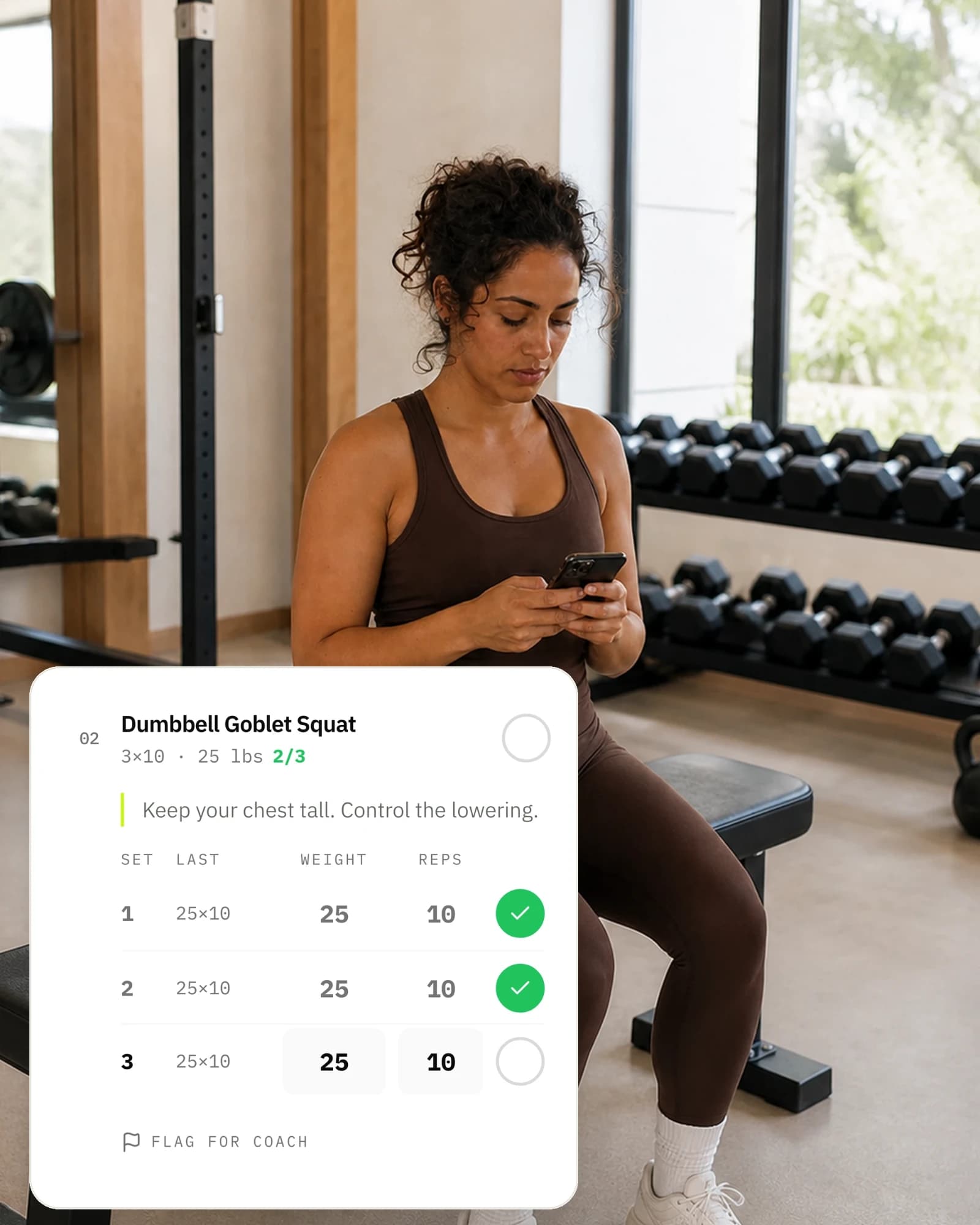 A client sitting on a gym bench between sets, logging a set on her phone: the Dumbbell Goblet Squat card shows two of three sets done and the coach's cue to keep the chest tall